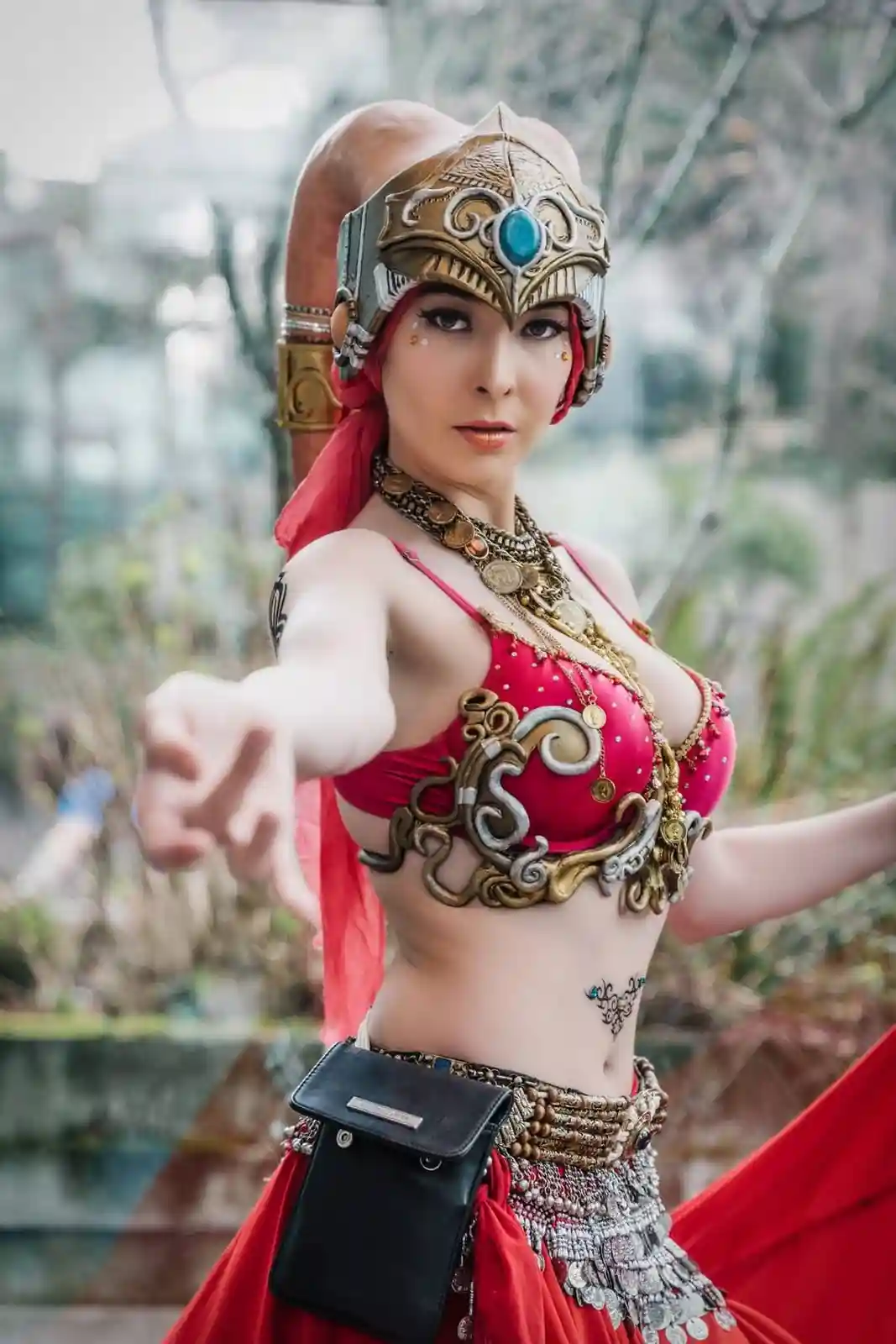 A woman in elaborate red and gold fantasy warrior cosplay, wearing a helmet, ornate top, and skirt, strikes a dramatic pose…