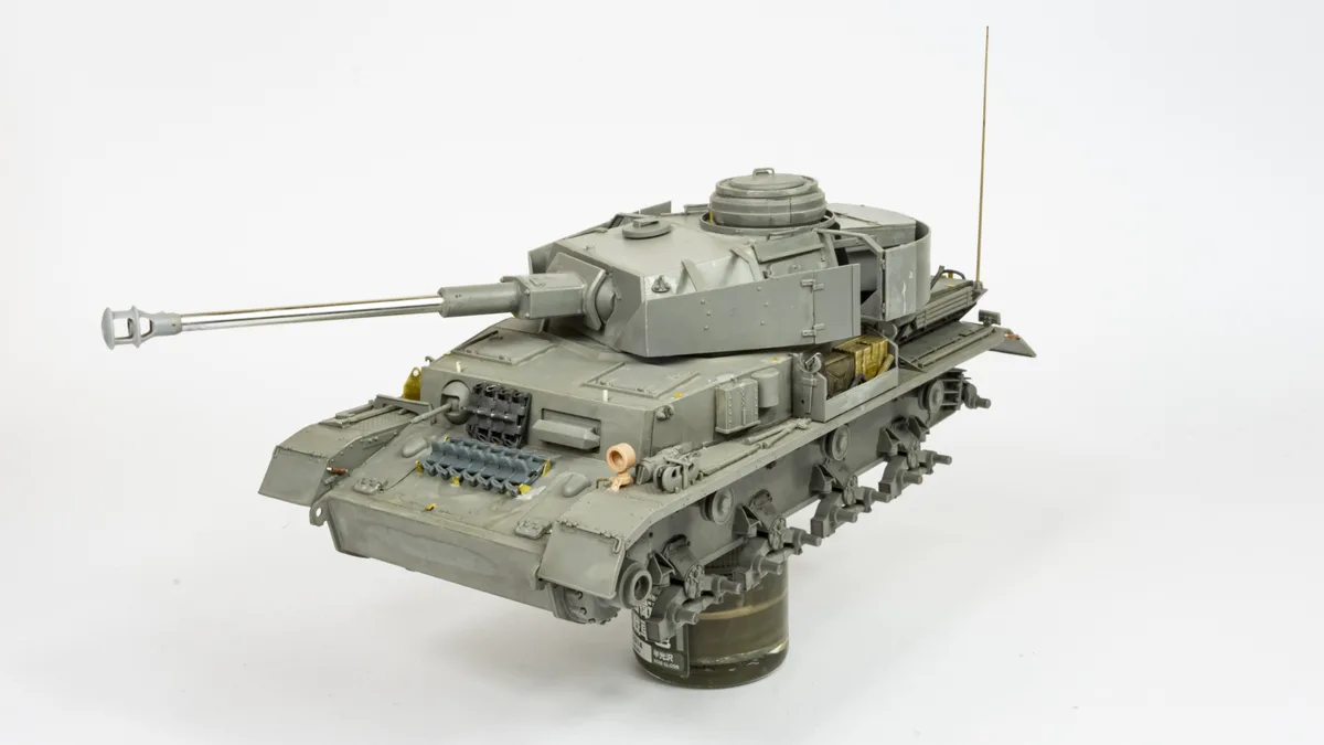 A detailed grey scale model of a tank is displayed on a cylindrical stand against a white background. The tank has a long…
