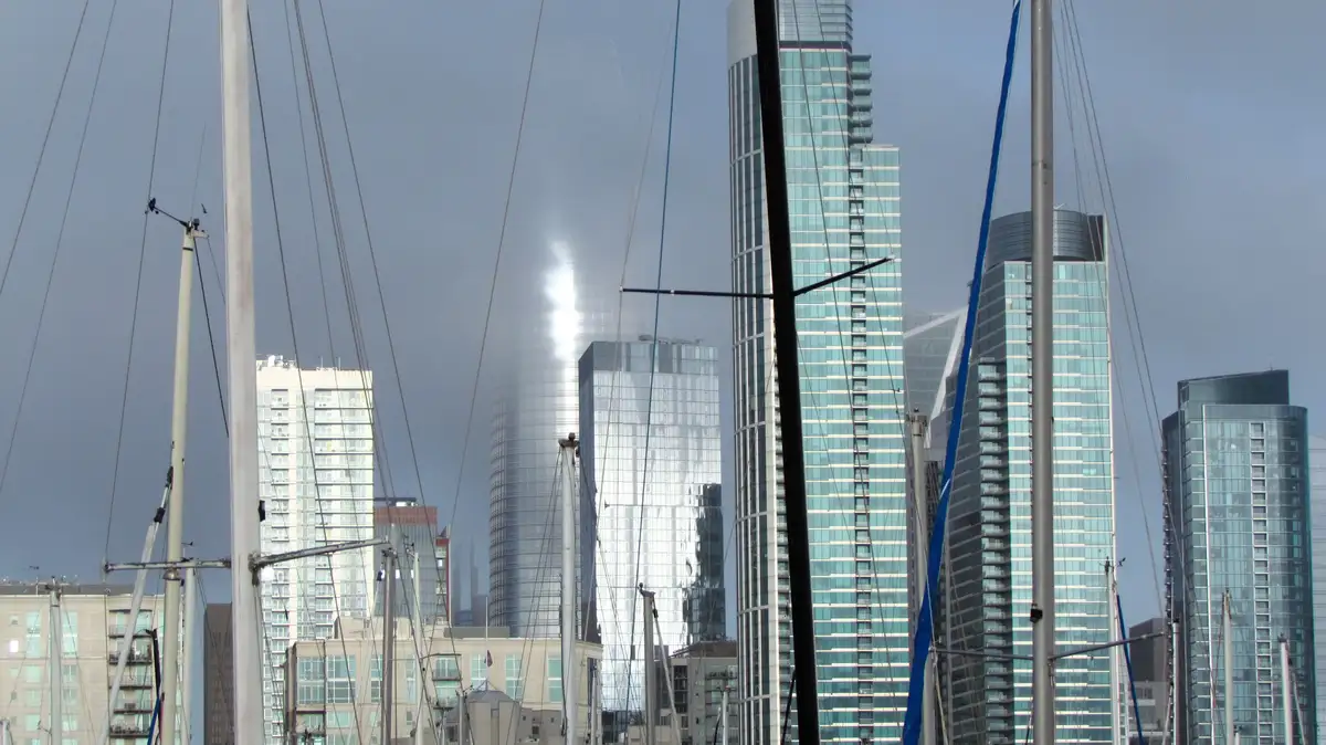 A city skyline with modern glass skyscrapers is viewed through the masts and rigging of sailboats in the foreground. The sun…