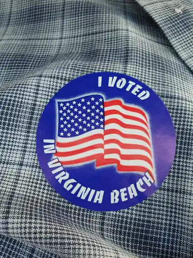 A close-up photo shows a circular blue sticker with a waving American flag design, worn on a blue and white checkered shirt…