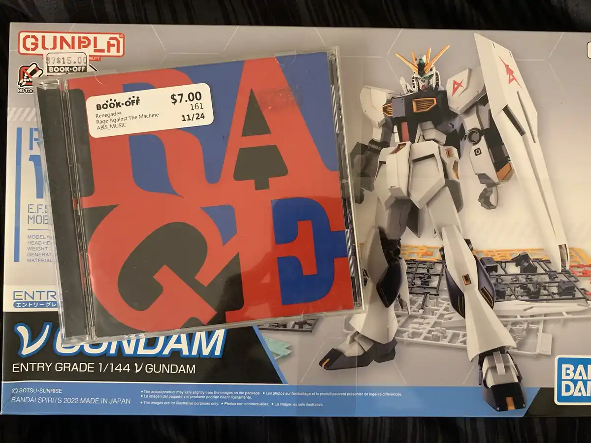 A $7.00 Book-Off price tag for a Rage Against The Machine CD is placed on top of a Bandai Gunpla model kit box for the 1/144…