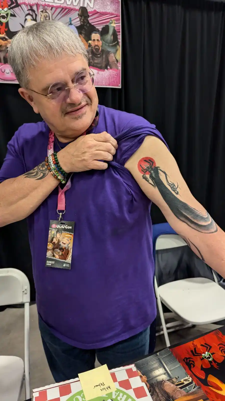 A man with gray hair and glasses, wearing a purple t-shirt and a convention lanyard, lifts his shirt to reveal a large…