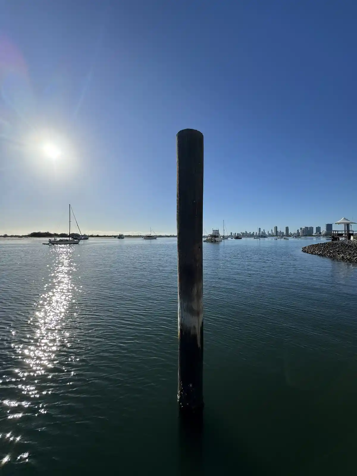 A tall, weathered wooden post stands upright in calm water under a bright sun, with a distant city skyline and boats visible…