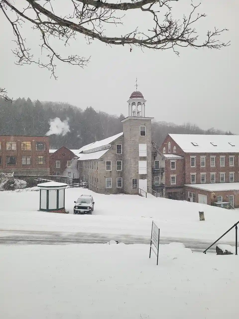 A snowy landscape shows a large, historic-looking stone and brick mill complex with a central tower, surrounded by…