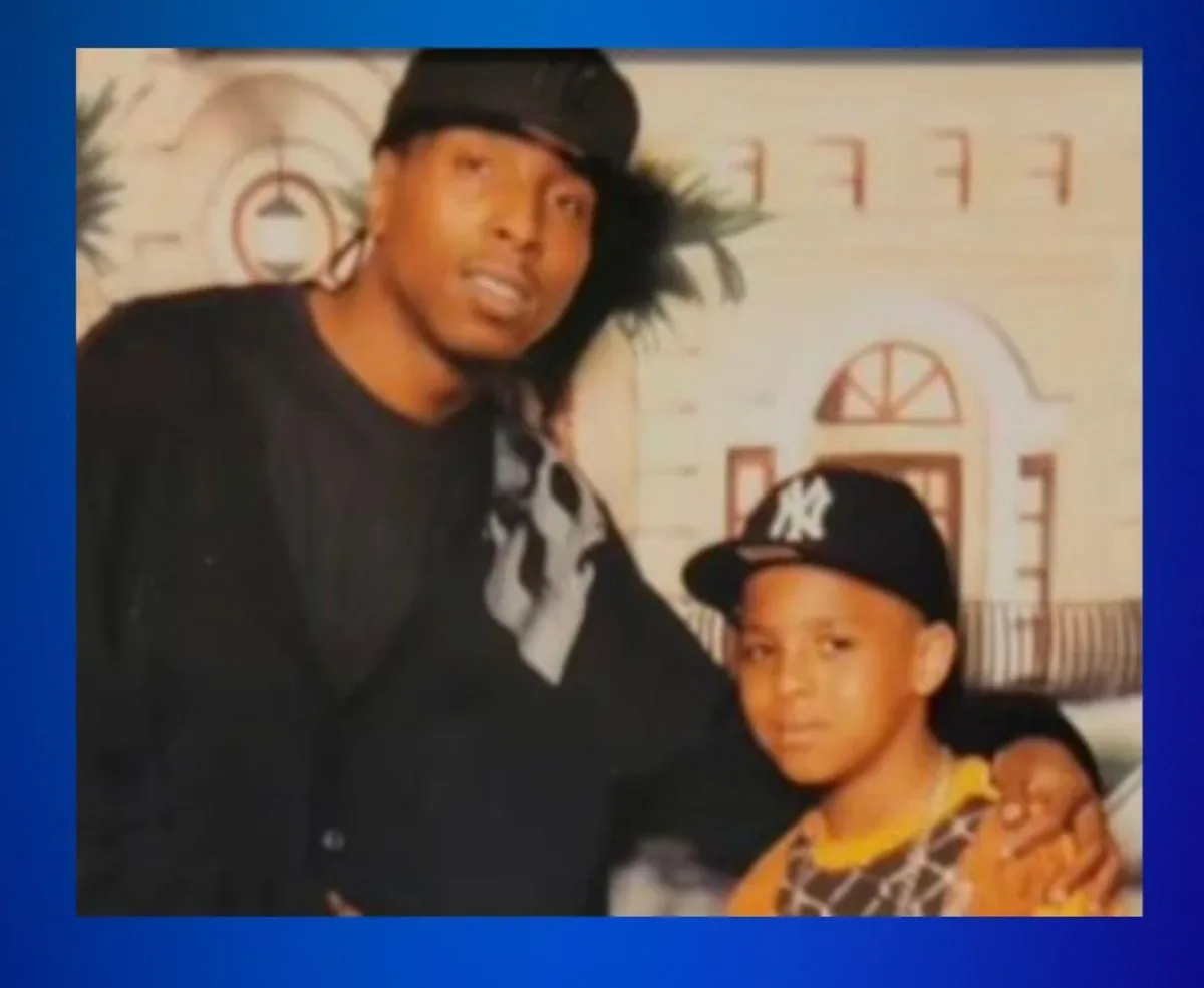 A grainy, old photograph shows two Black men posing together; an adult man in a black cap and jacket has his arm around a…