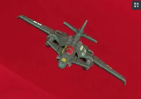 A dark green toy fighter jet with "ALLAH" written in yellow on its side is shown against a solid red background. The jet has…