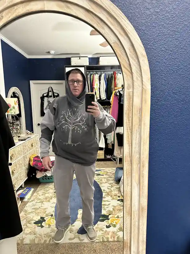 A man wearing a grey hoodie with a white tree design on the front stands in front of a large arched mirror, holding a phone…