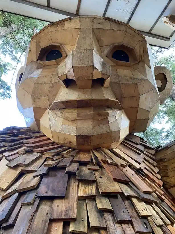 A low-angle photo shows a giant, geometrically faceted wooden sculpture of a human face with dark, hexagonal eye sockets and…
