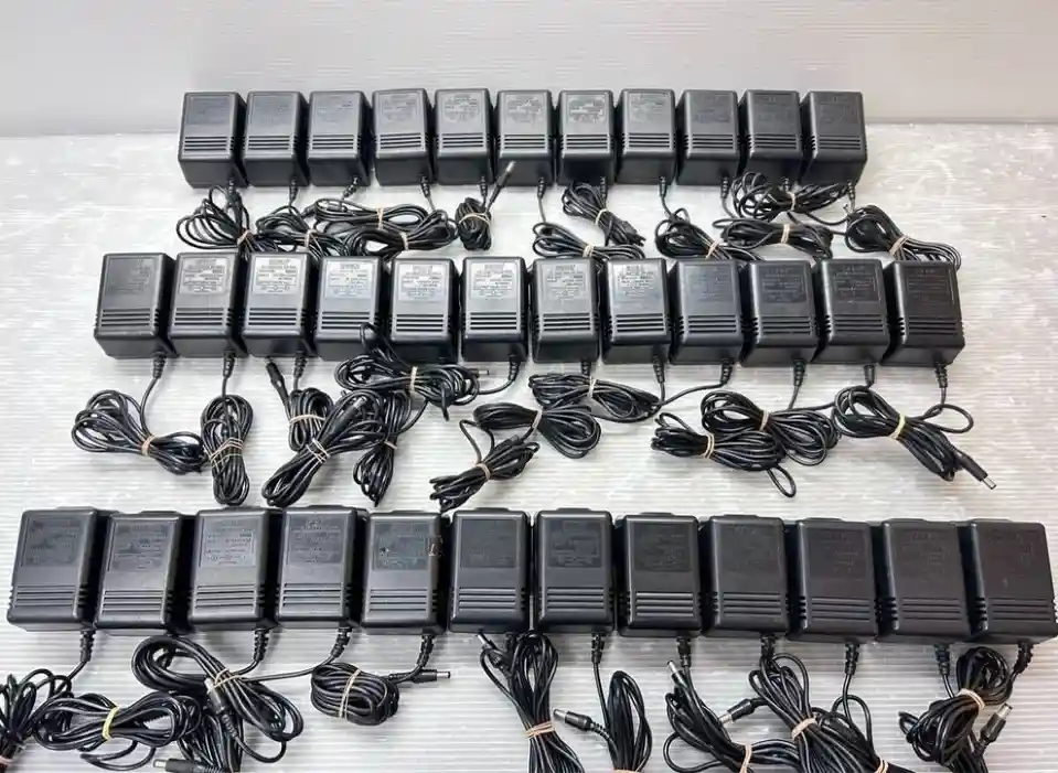 An overhead shot displays dozens of identical black AC adapters arranged in neat rows on a light gray surface. Each adapter…