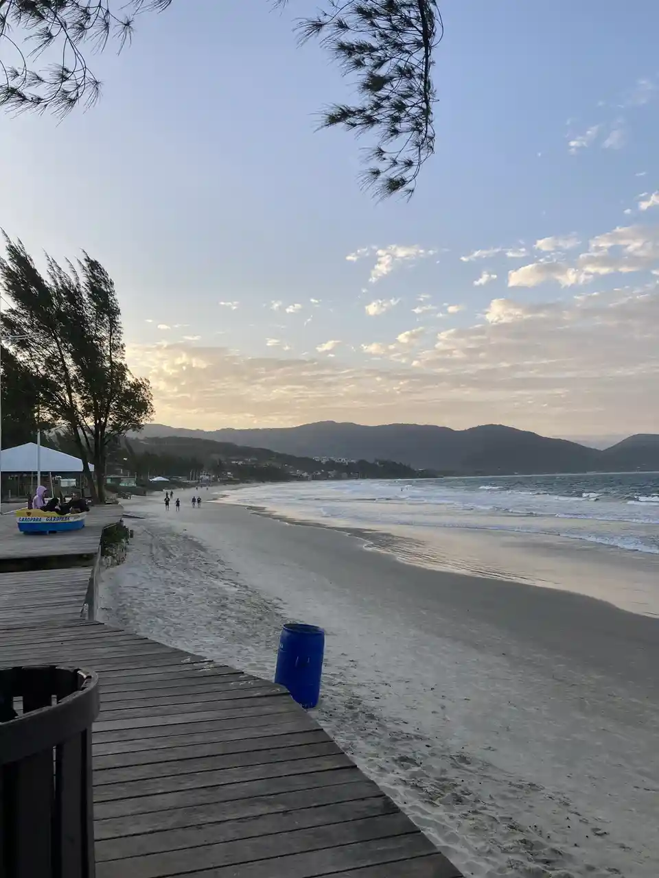 A serene beach scene at dawn or dusk with a wooden boardwalk in the foreground, a blue barrel on the sand, and distant hills…