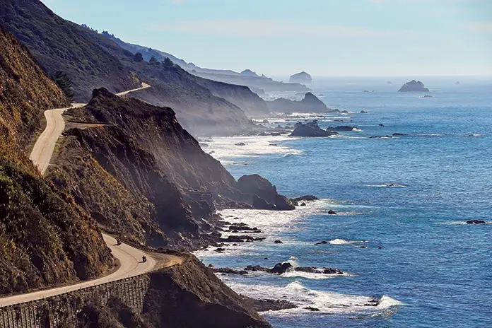 A scenic photo of a winding coastal road along a steep cliffside overlooking the ocean, with a few small vehicles visible on…
