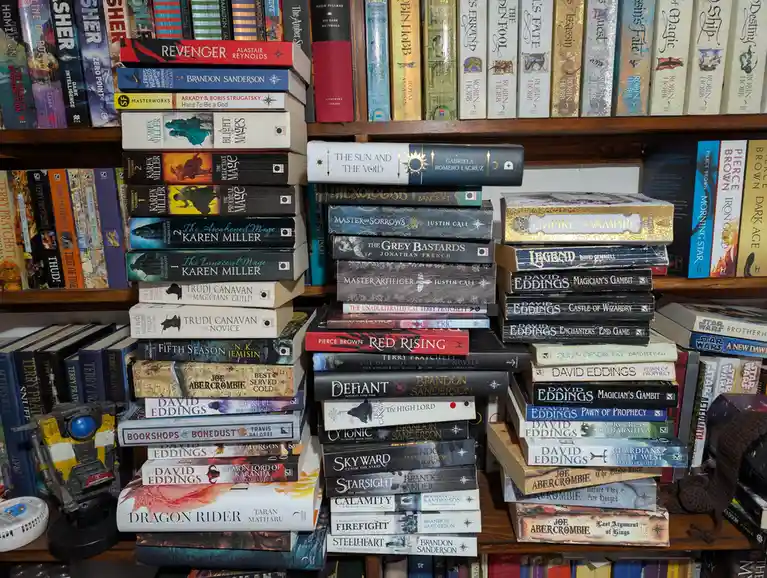 A wooden bookshelf is densely packed with stacks of fantasy and sci-fi novels by authors like Brandon Sanderson, David…