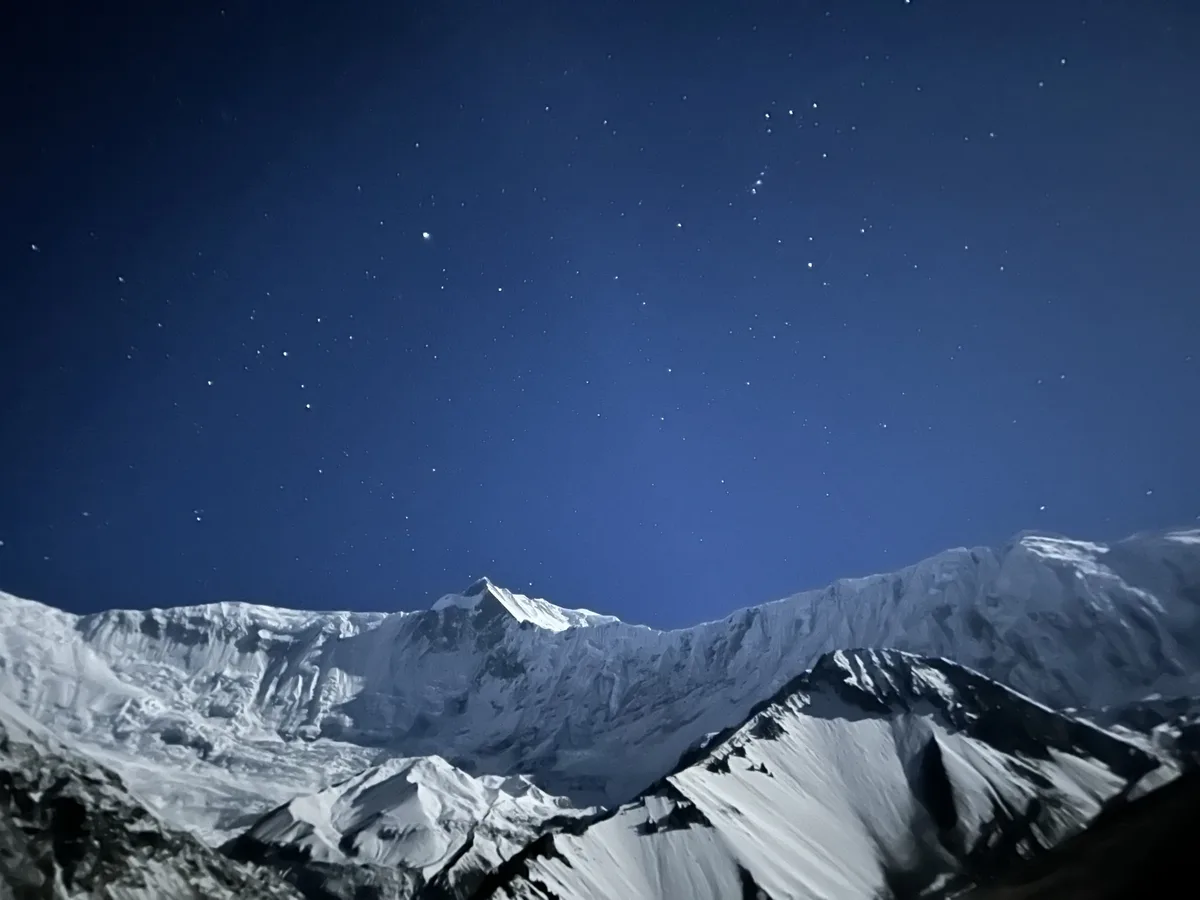 A breathtaking night photograph shows snow-capped mountain peaks under a deep blue sky filled with countless stars. The…