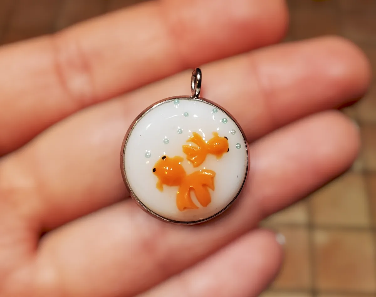 A close-up photo shows a hand holding a small, round, silver-framed pendant. Inside the pendant, two stylized orange…