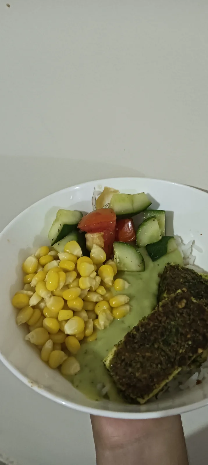 A hand holds a white bowl containing a healthy meal with rice, yellow corn kernels, chopped cucumber, diced tomato, a green…