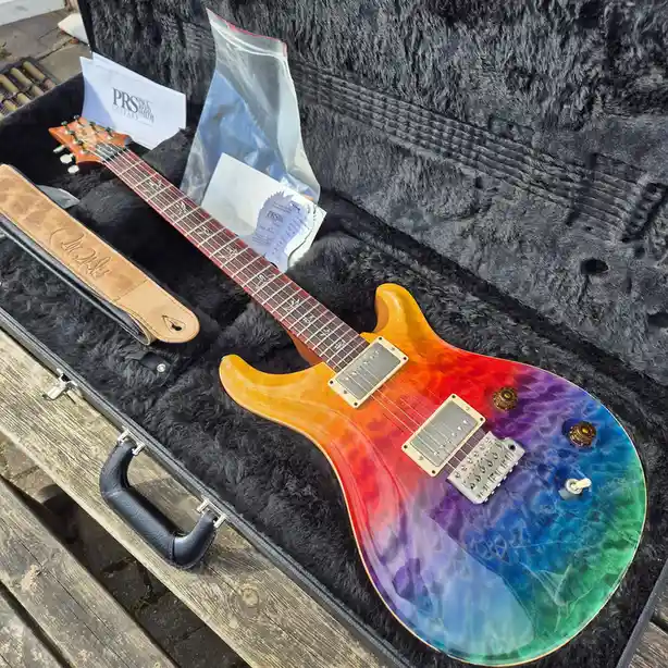 A PRS electric guitar with a vibrant rainbow finish rests inside its black, fuzzy-lined case, which also contains a branded…