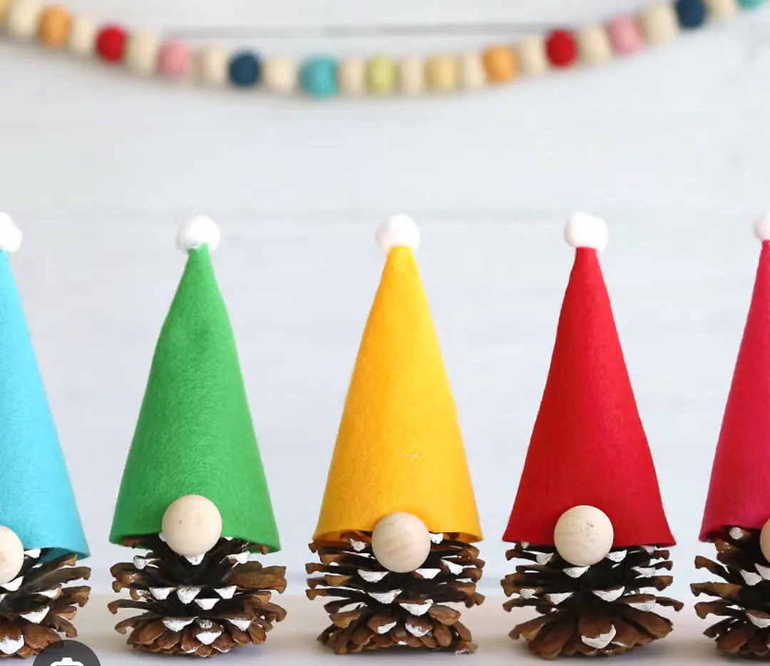 A row of handmade gnomes made from pinecones with felt hats in various colors (blue, green, yellow, red) and white pom-pom…