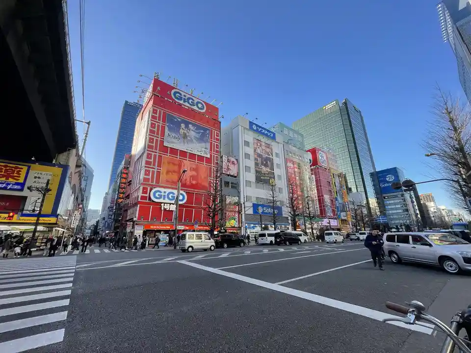 A wide-angle photo of a busy Tokyo street intersection under a clear blue sky, featuring tall buildings covered in large…
