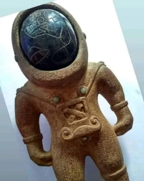 A small, brown, textured figurine of an astronaut with a cracked, dark blue globe inside its helmet, standing on a white…
