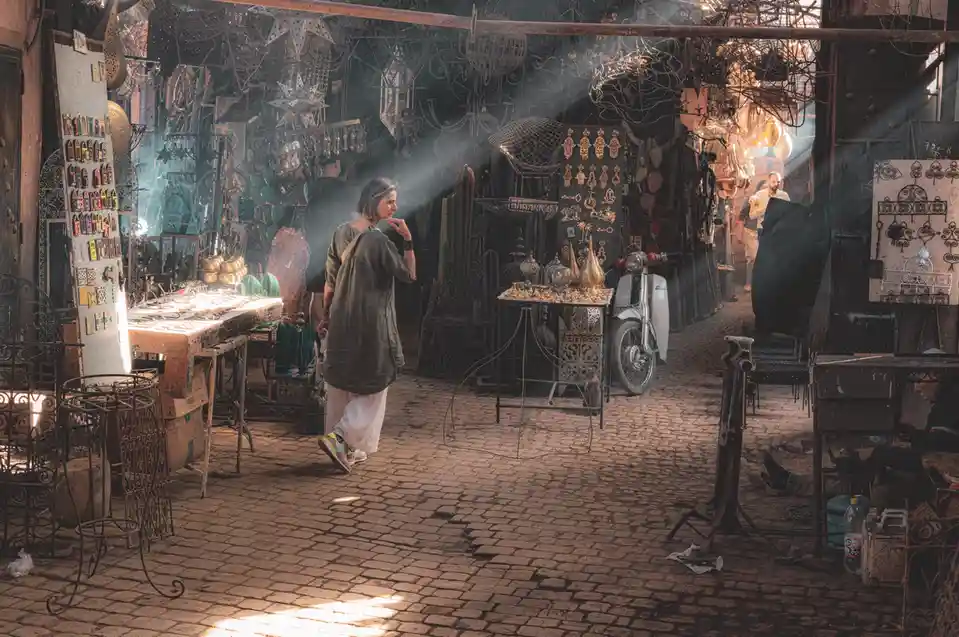 A woman walks through a dim, atmospheric market stall filled with ornate metalwork, hanging lanterns, and jewelry displays…