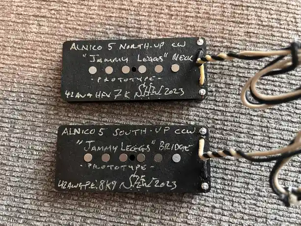 Two custom guitar pickups lie on a textured surface, each with handwritten labels. The top pickup is labeled 'ALNICO 5…