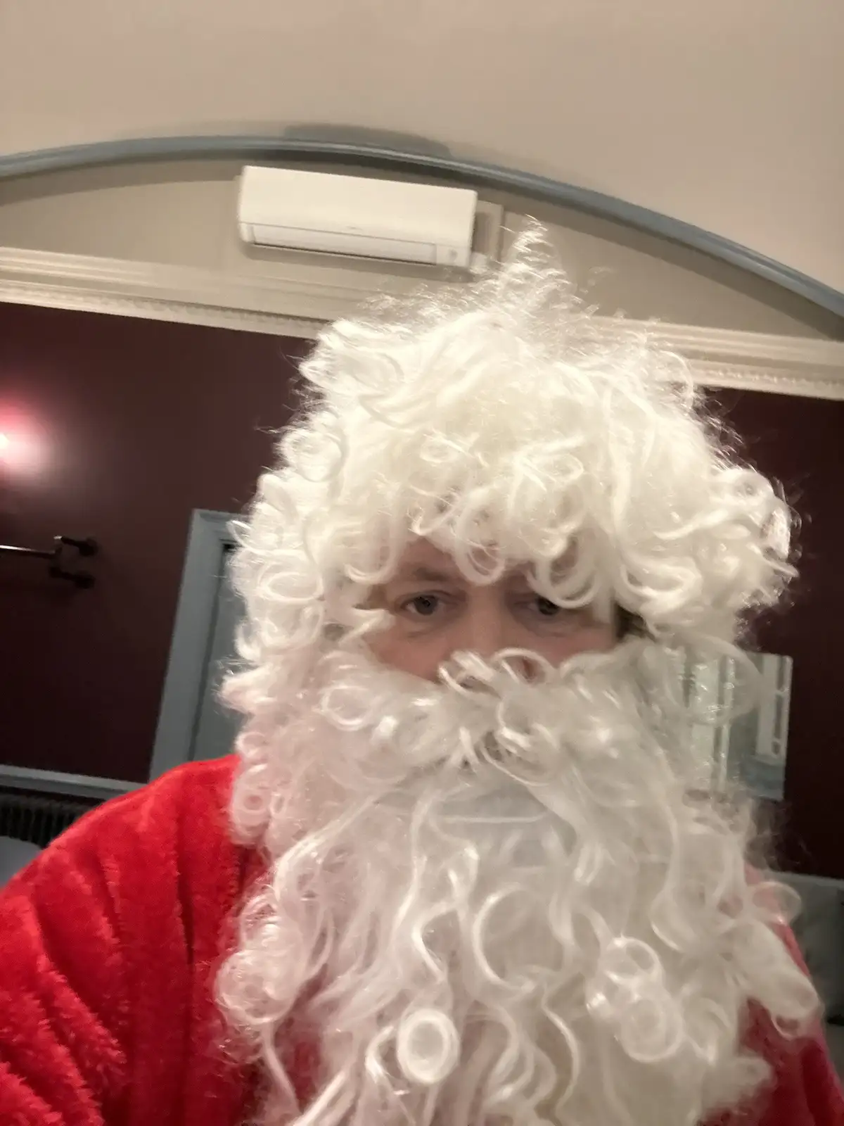 A person wearing a full Santa Claus costume, including a red robe and a large, curly white wig and beard, looks directly at…