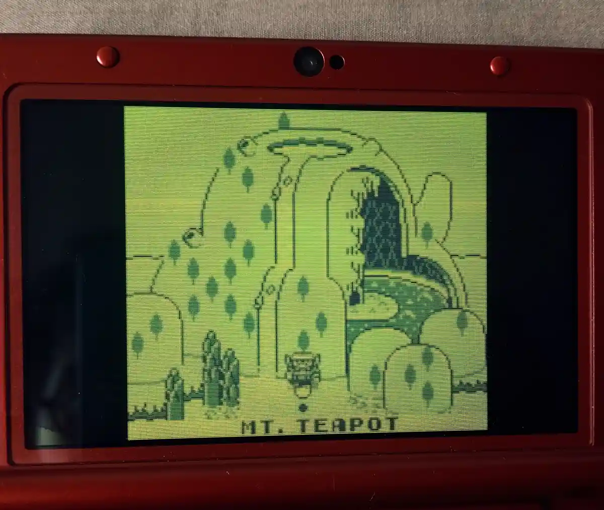 A red Nintendo DS displays a pixelated, green-tinted map from a game, showing a mountainous area with trees and a small…