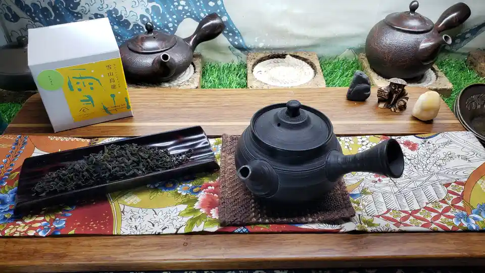 A still life of a traditional tea setup with a black ceramic teapot on a woven mat, a tray of loose-leaf tea, a tea box, and…