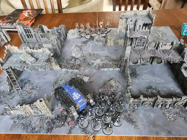An overhead view of a Warhammer 40,000 tabletop battle scene on a wooden table, featuring miniature figures and terrain…