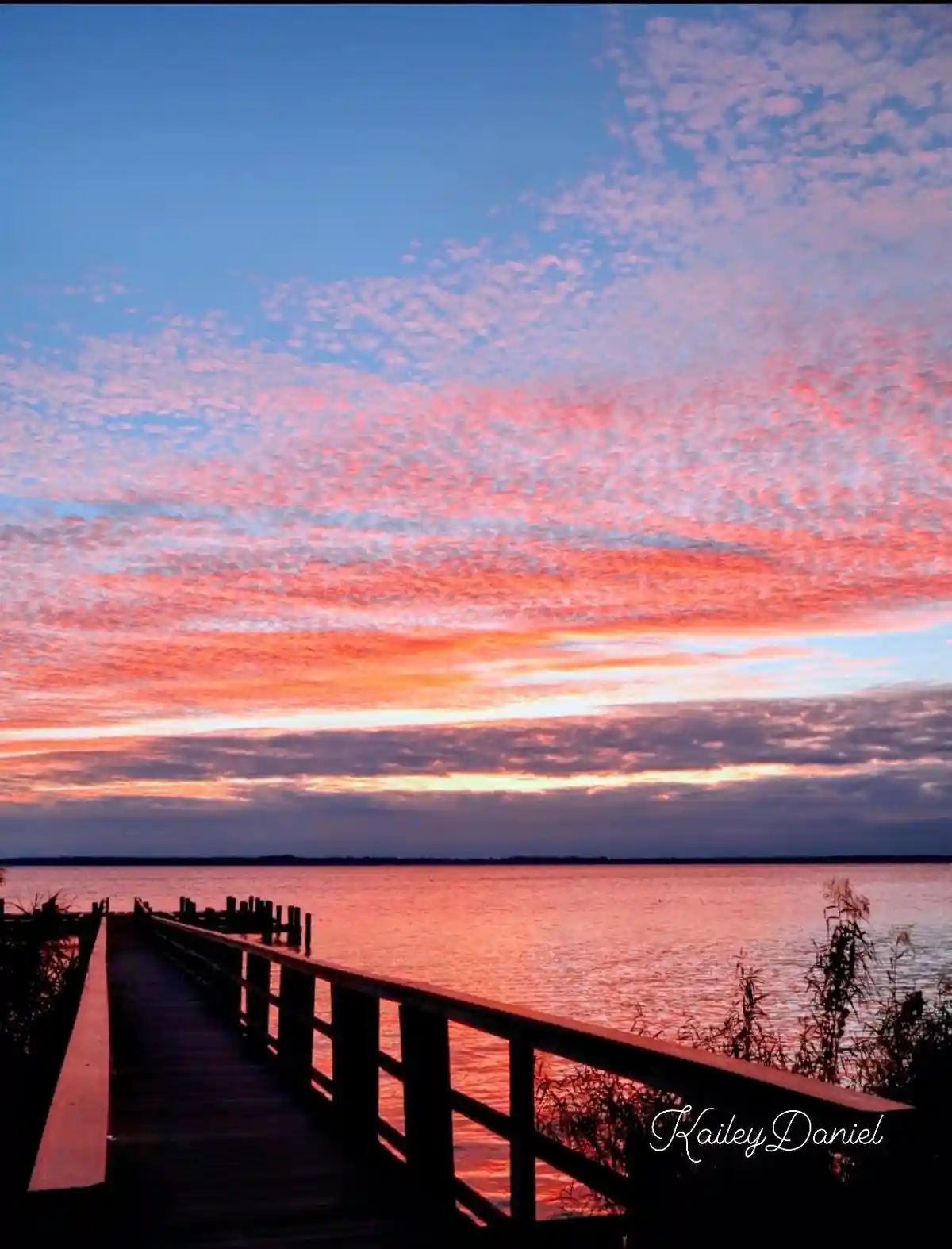 A serene sunset photo shows a wooden pier extending into a calm body of water, with clouds painted in vibrant pink, orange…
