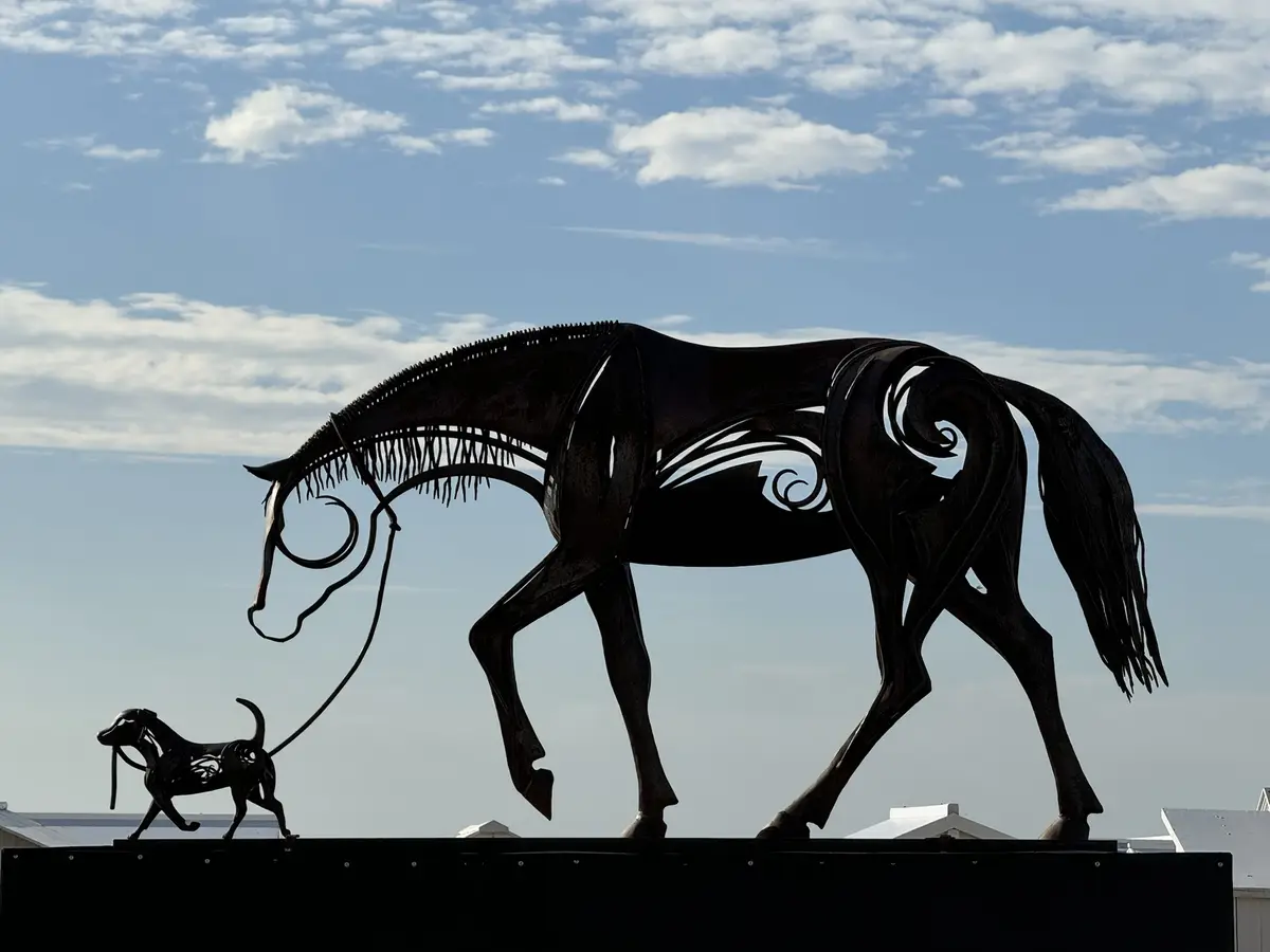 A metal sculpture of a large horse and a small dog, both in silhouette, with the dog holding a leash that extends to the…