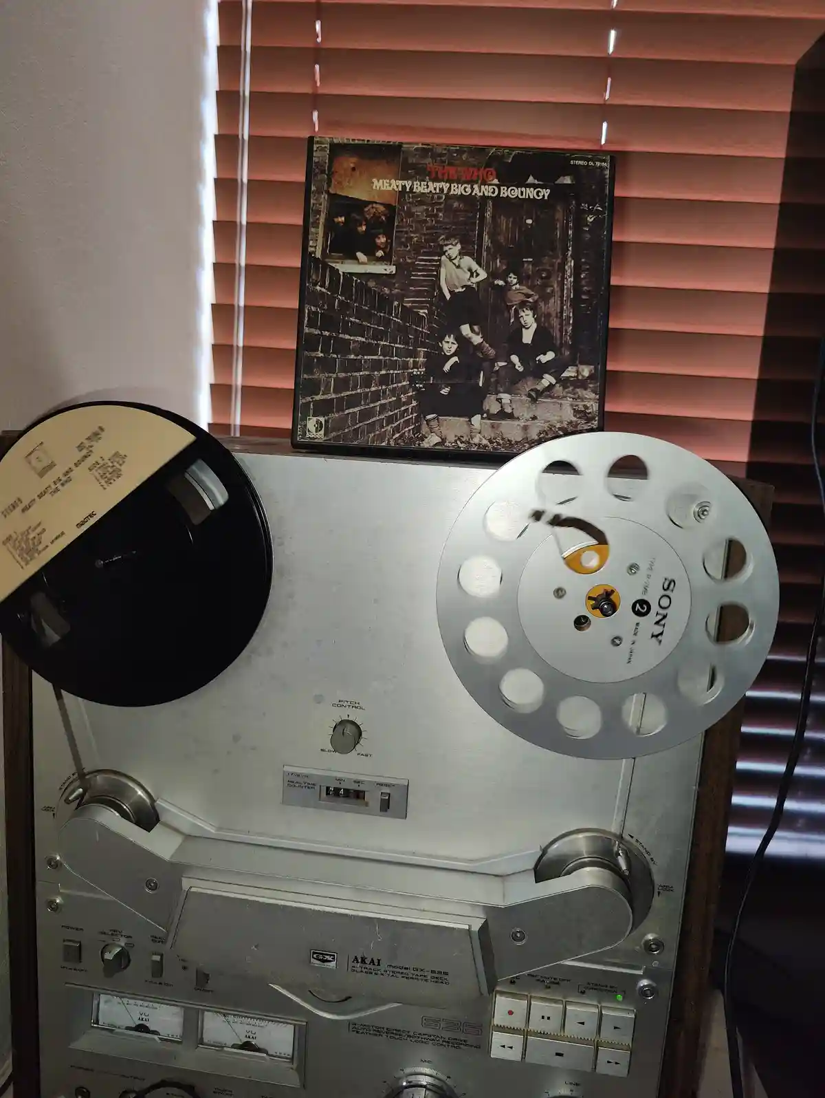 A vintage Akai reel-to-reel tape recorder with a black and silver spool setup sits in front of a window with blinds. On top…