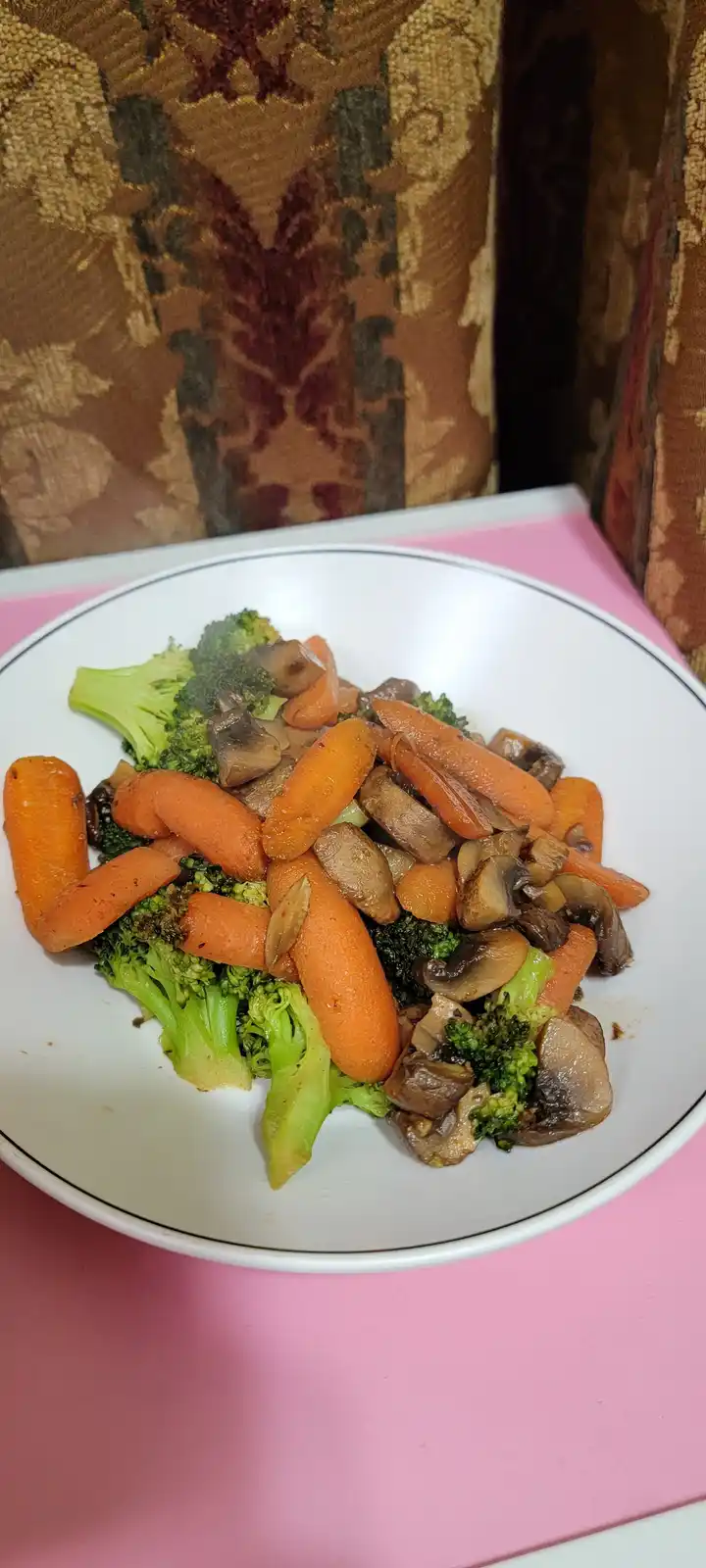 A white plate with a thin blue rim holds a serving of cooked vegetables — bright green broccoli florets, orange baby…