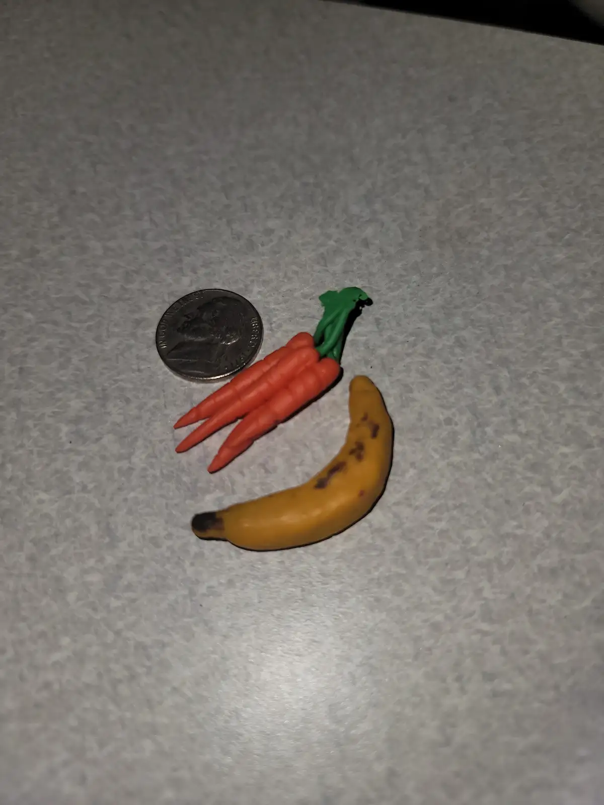 A close-up photo shows a tiny plastic toy banana and a toy carrot bundle next to a U.S. quarter on a speckled gray surface…