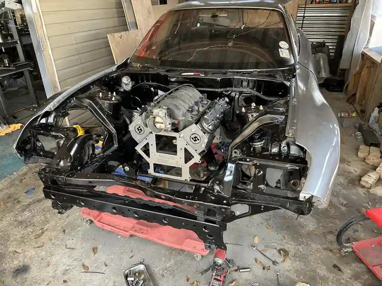 A silver classic car is shown in a garage with its hood open, revealing a partially assembled engine. The engine block has…