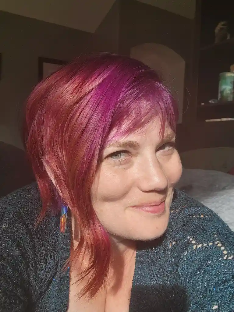 A close-up photo shows a woman with short, vibrant magenta and reddish-pink hair, wearing a sparkly dark sweater and a…