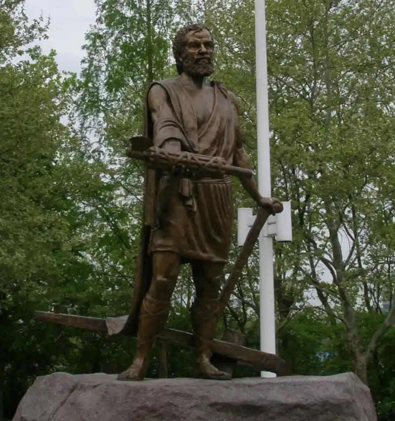 A bronze statue of a bearded man in ancient-style robes stands on a stone pedestal, holding a large wooden crossbar. A…