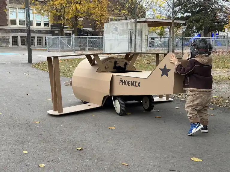 A young child wearing a pilot helmet and jacket stands beside a large, homemade cardboard airplane with wheels, labeled 'THE…