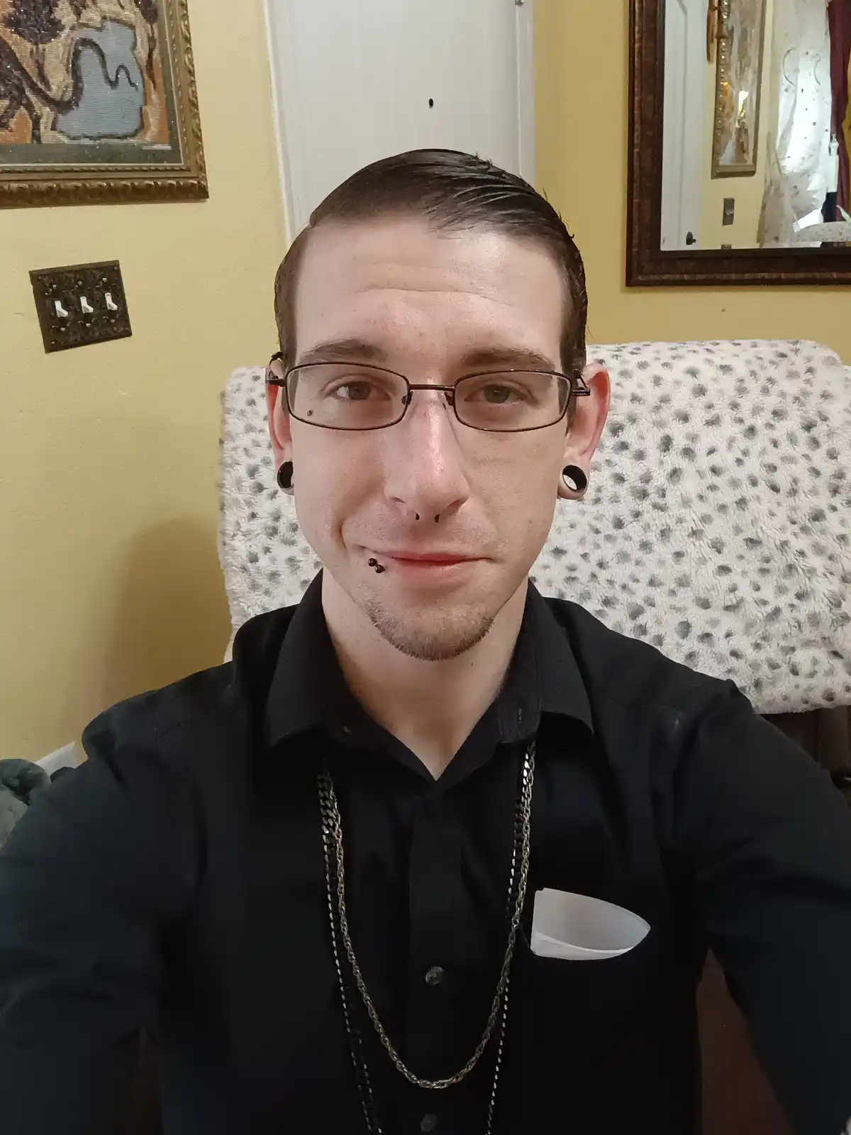 A man with glasses, multiple facial piercings, and black ear gauges sits in a chair wearing a black shirt with a white…