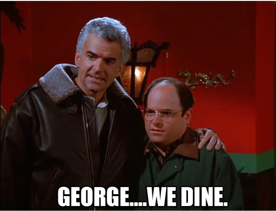 Two men stand together in a red-walled room; the taller man in a leather jacket has his arm around the shorter man in a…
