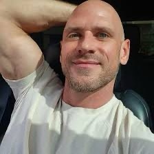 A bald man with a light beard, wearing a white t-shirt, sits in a car seat and smiles at the camera while resting his right…