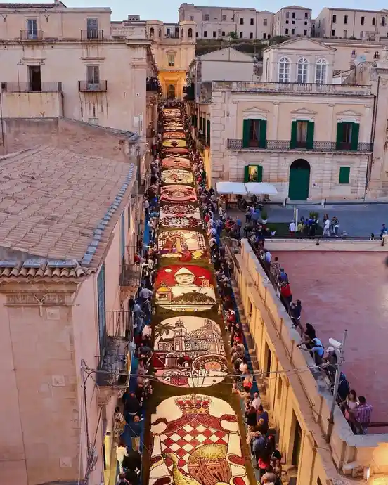 An aerial view shows a narrow street in a European town filled with people watching a long, intricate floral carpet laid out…