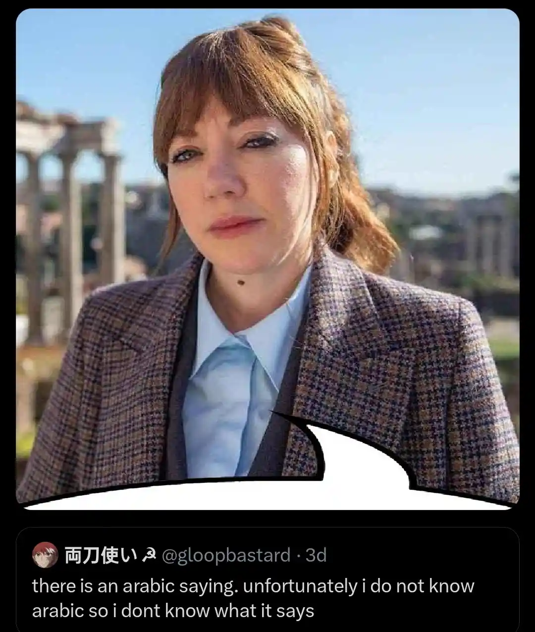 A woman with reddish-brown hair in a half-updo, wearing a patterned blazer over a light blue collared shirt and tie, looks…
