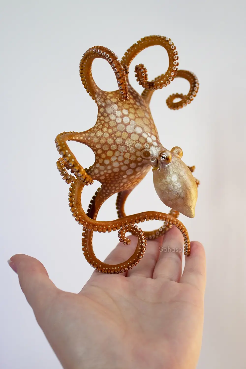 A small, intricately detailed golden-brown octopus sculpture with white spots and curled tentacles is balanced on the palm…