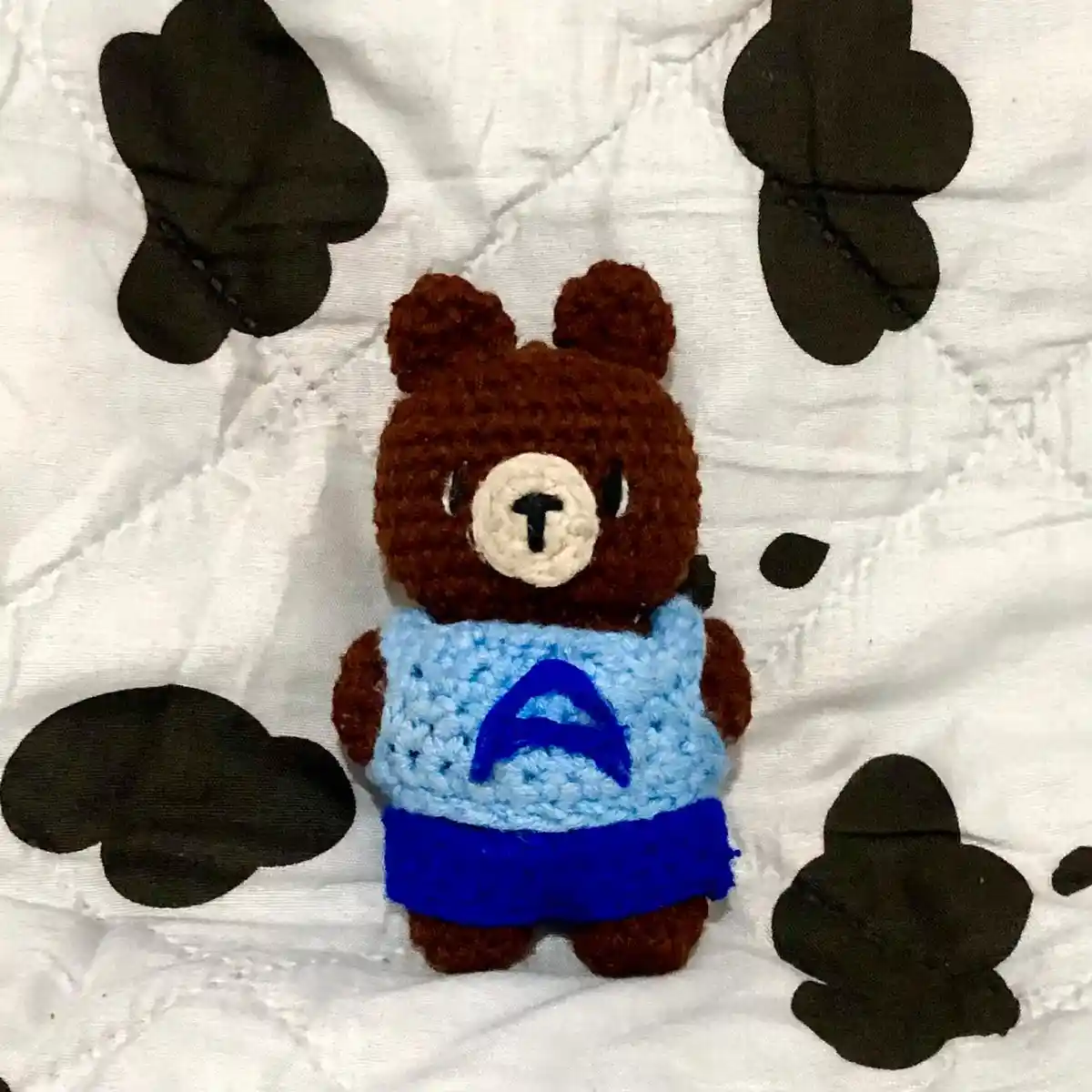 A handmade crocheted brown bear plushie wearing a light blue vest with a large blue letter 'A' on the front and blue shorts…