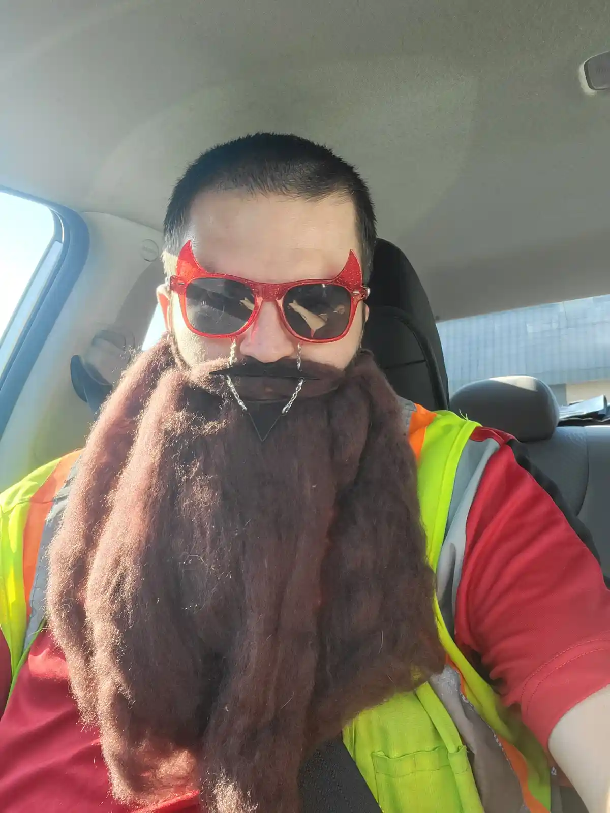 A man in a car wearing a bright yellow safety vest over a red shirt, sporting a long brown fake beard, red glittery…