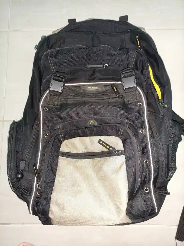A black Targus backpack with multiple zippers and compartments lies open on a tiled floor, revealing an empty beige front…