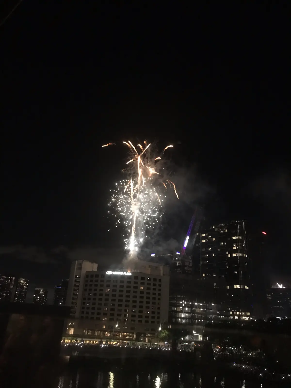 A nighttime cityscape with fireworks exploding above a building labeled 'CROWN PLAZA', with a crane and other skyscrapers…