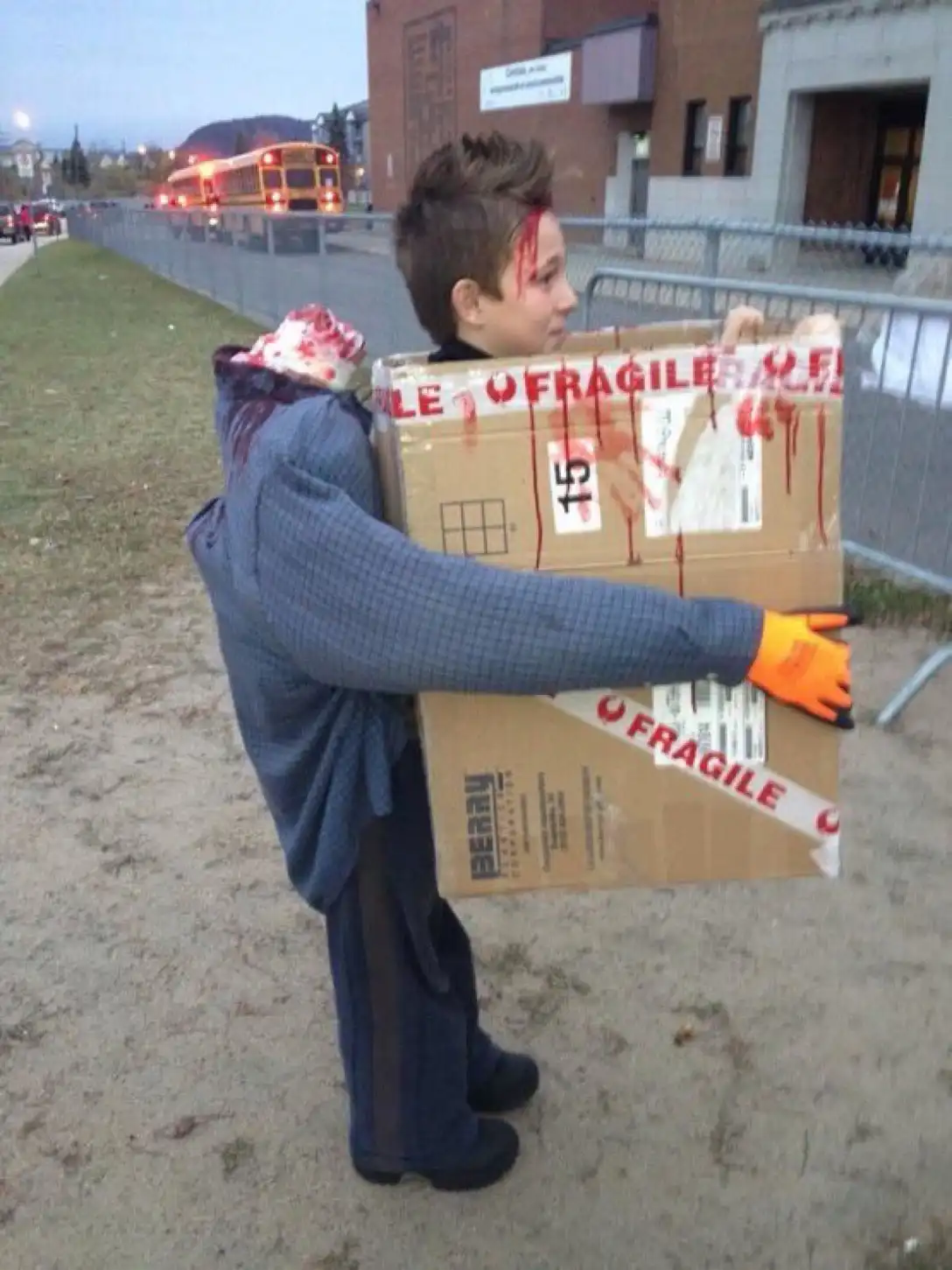 A young boy dressed as a zombie with fake blood on his face and neck, carrying a large cardboard box labeled 'FRAGILE' with…