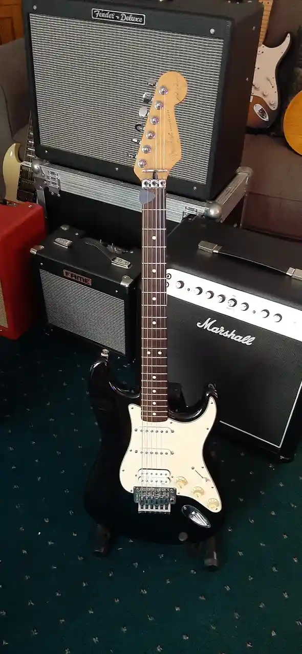 A black Fender Stratocaster electric guitar stands upright on a stand in front of several amplifiers, including a Fender…